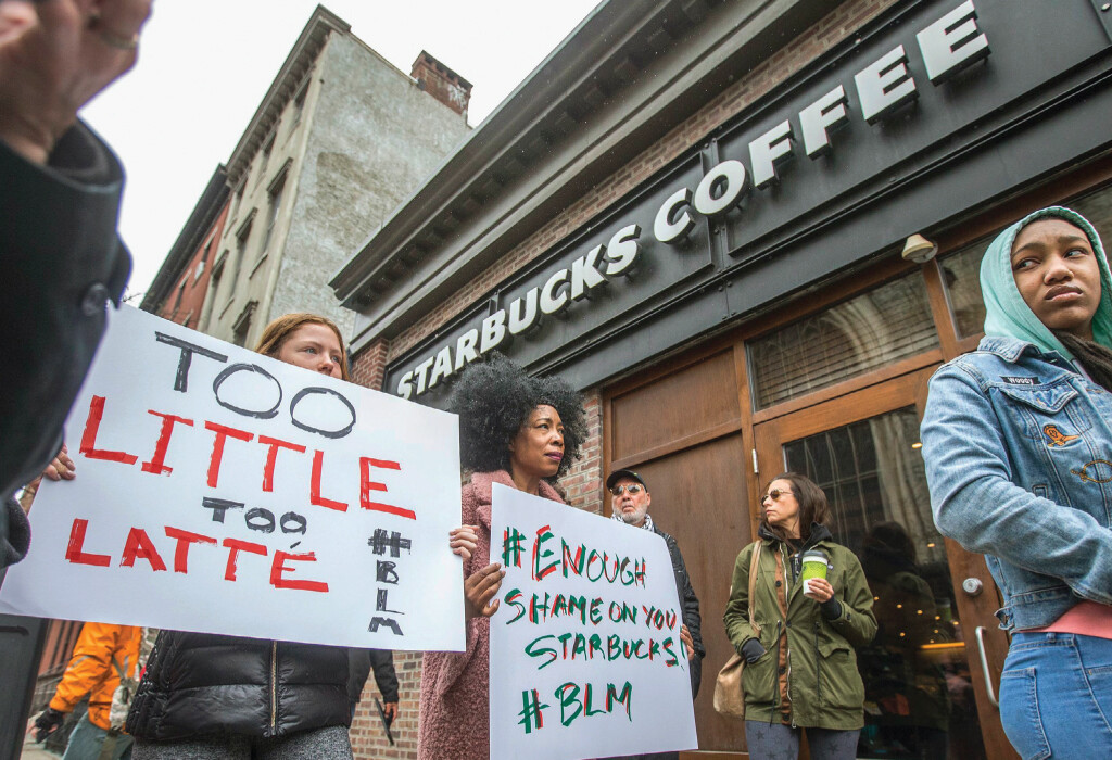 Protestors stand in front of a Starbucks Coffee shop, two of which hold up posters.
