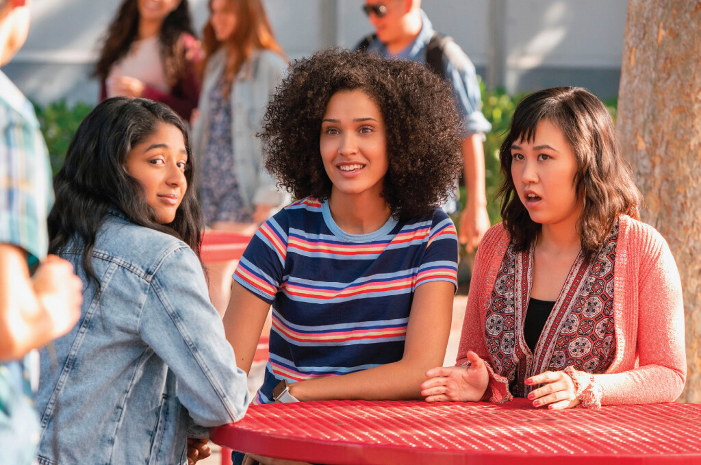 A scene from Never Have I Ever features three high school-aged girls of color sitting at an outside picnic table.