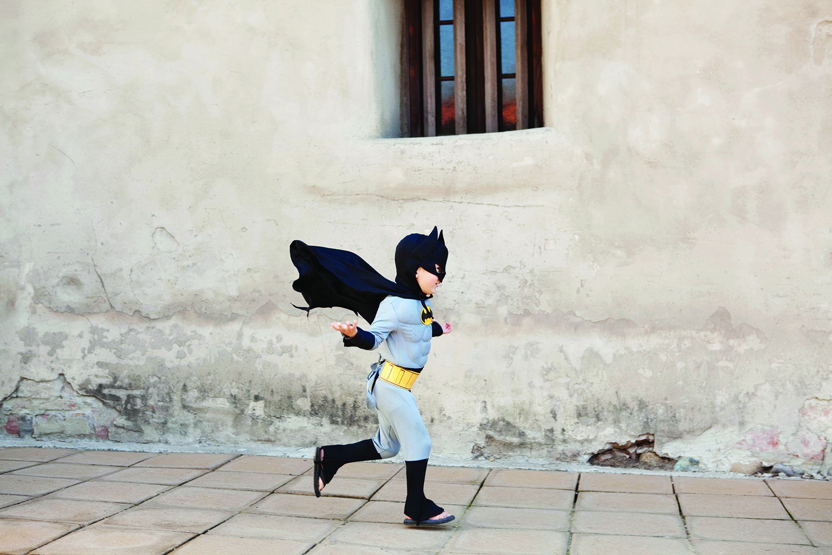A kid runs down the street wearing a batman costume.