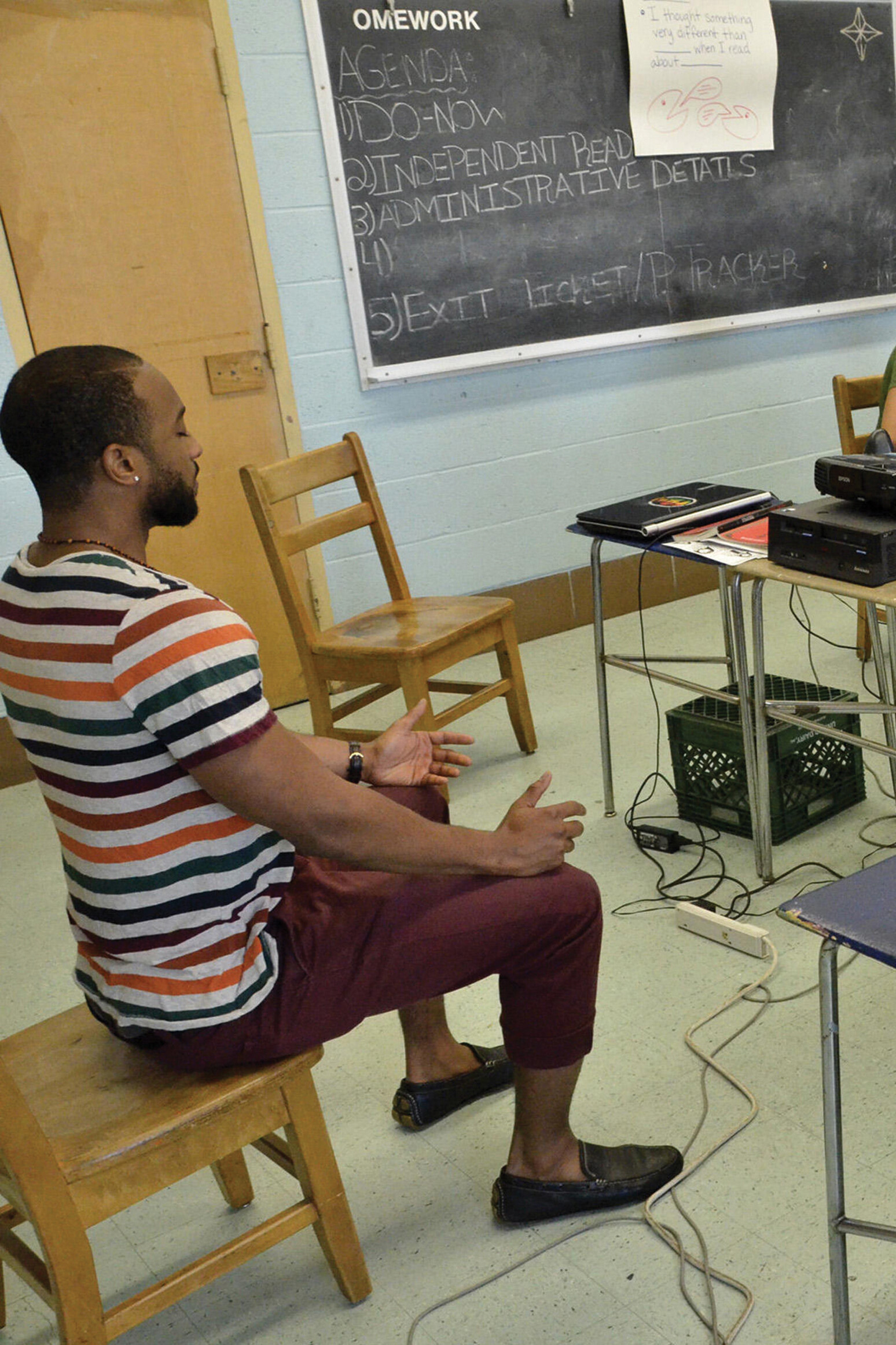 A man sits on the edge of his seat with perfect posture in a classroom. His arms rest on his knees, and his eyes are closed.