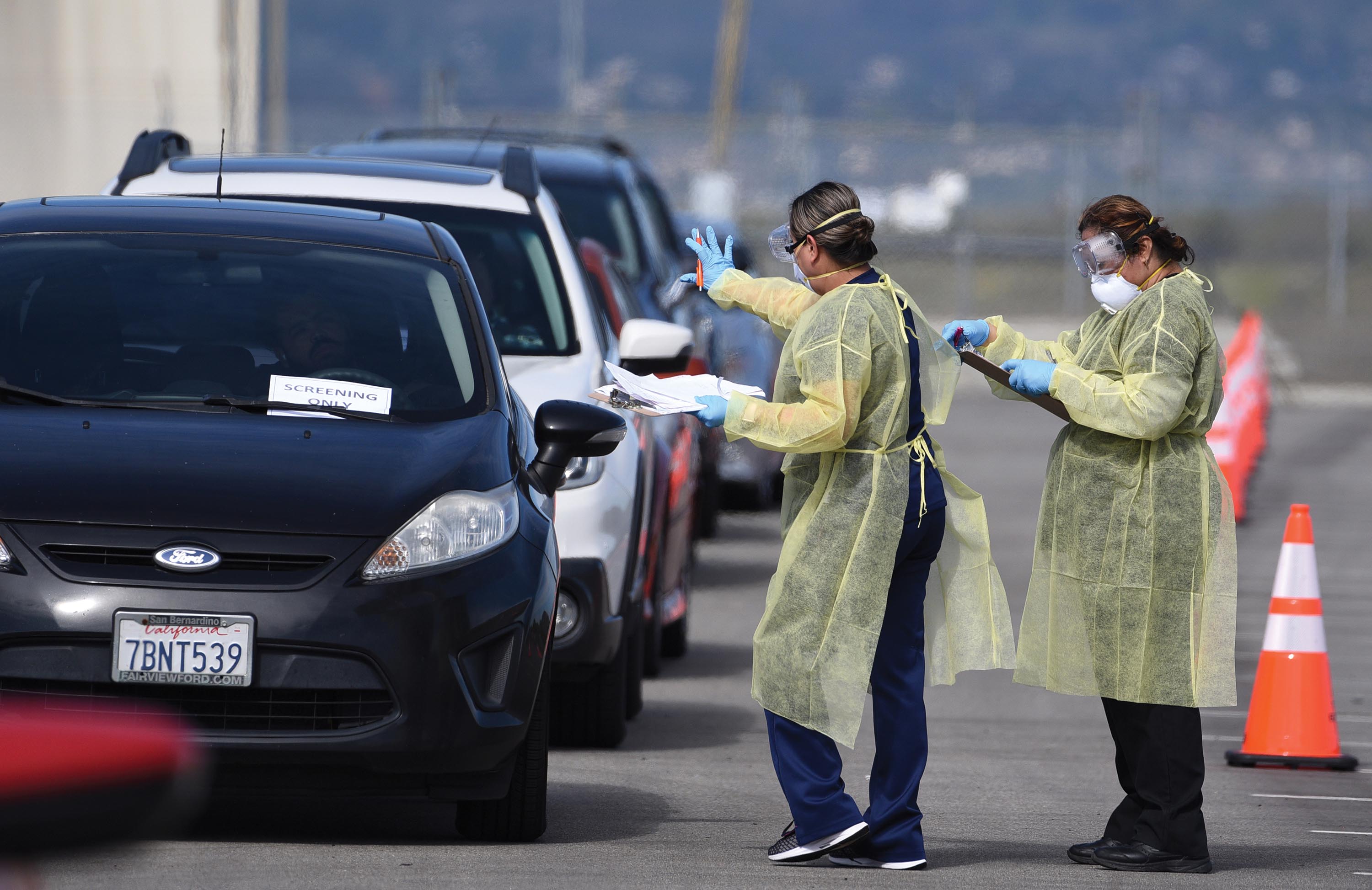 Two women wearing gloves and aprons standing on a highway. One of the women gestures toward a row of cars.