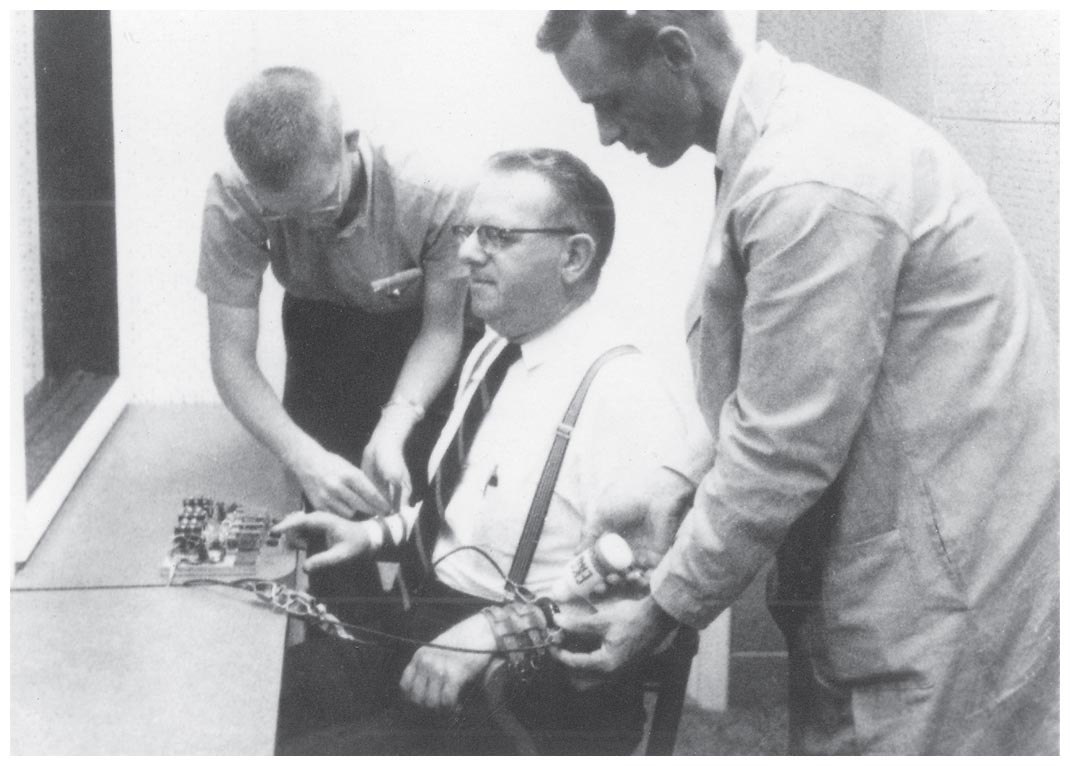 Two men attach electrodes on the hands of a third person who sits on a chair.