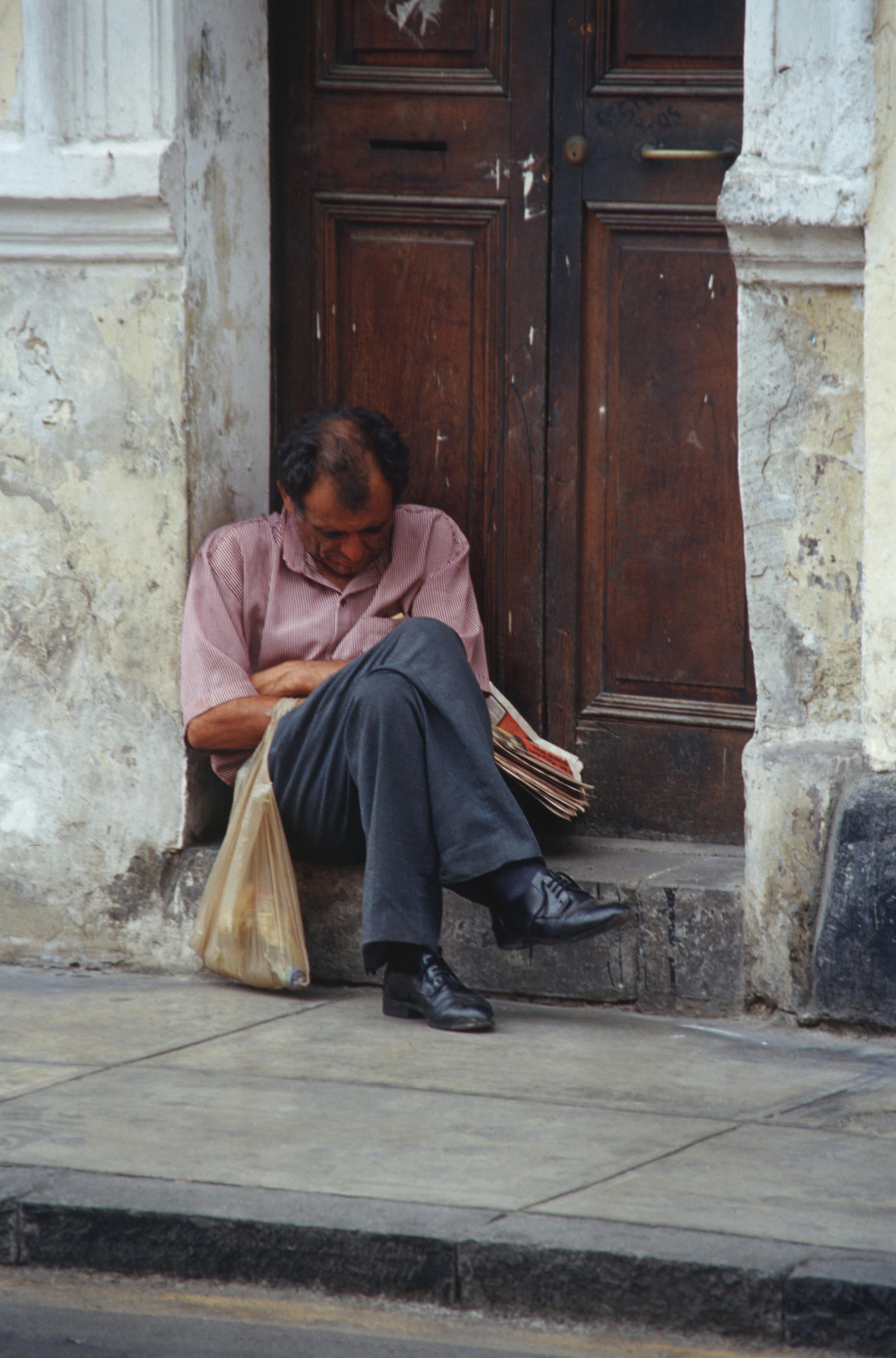 A man sitting at the threshold of a closed door with his head legs crossed and head bent down. He is carrying a bag and a stack of papers.