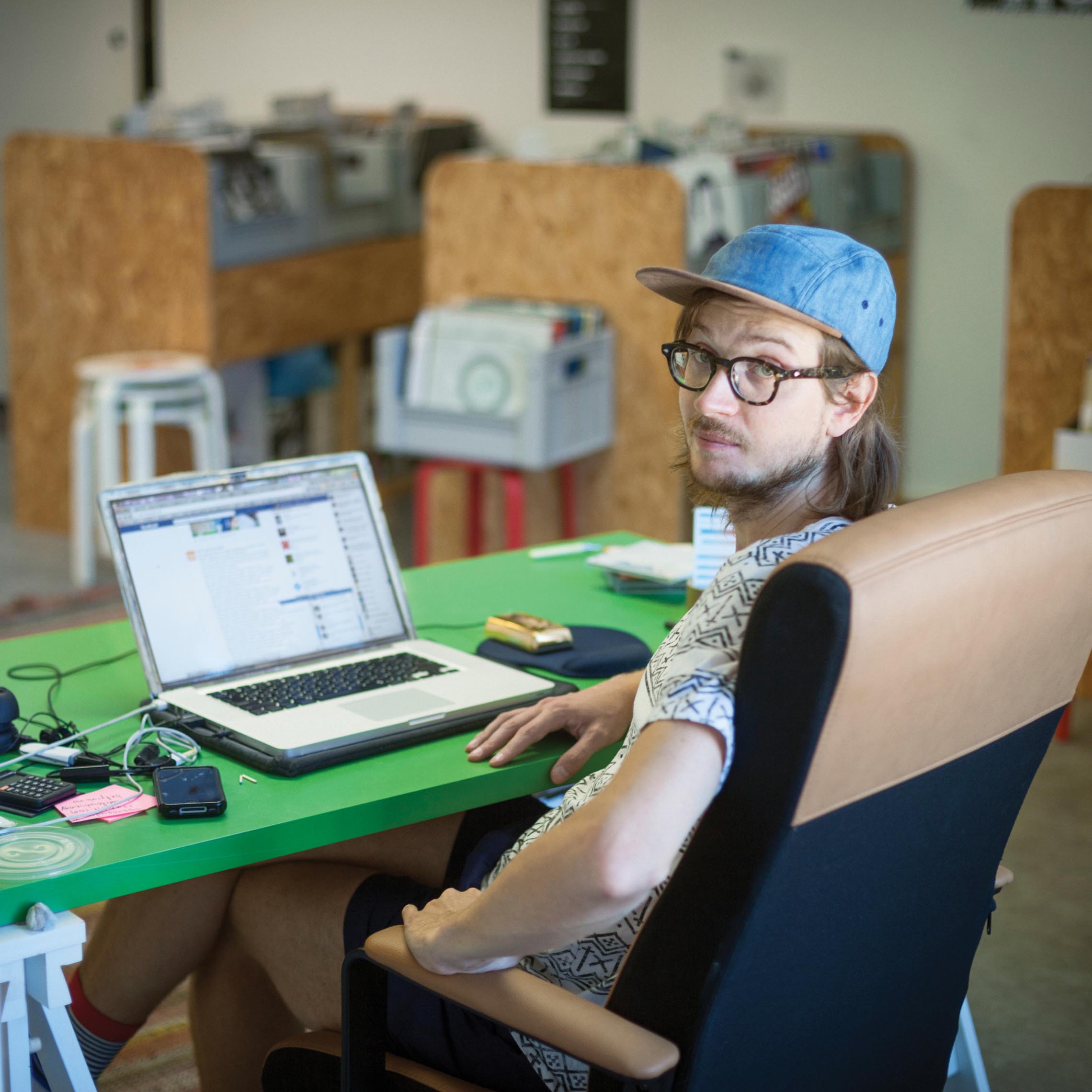 A bespectacled young man wearing a cap and a t-shirt sitting in front of his laptop.