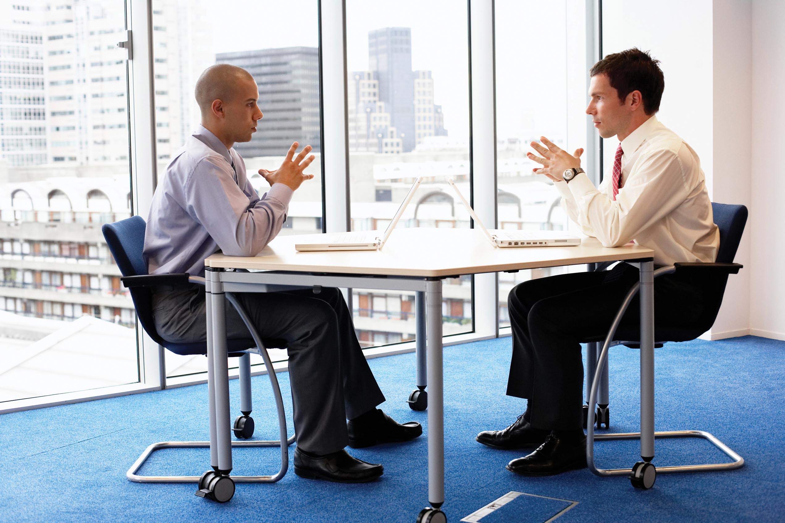 Two men sit facing each other. Both of them rest their elbows on the table with their fingers touching each other.