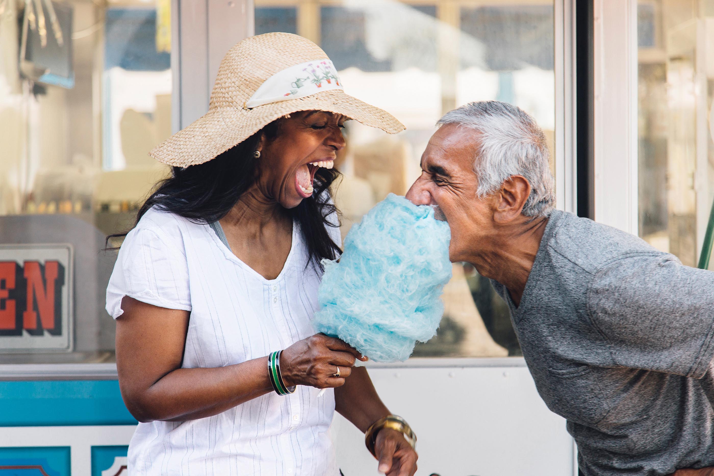 A laughing woman holds a cotton candy in her hand. An elderly man bends forward and takes a bite of the candy.