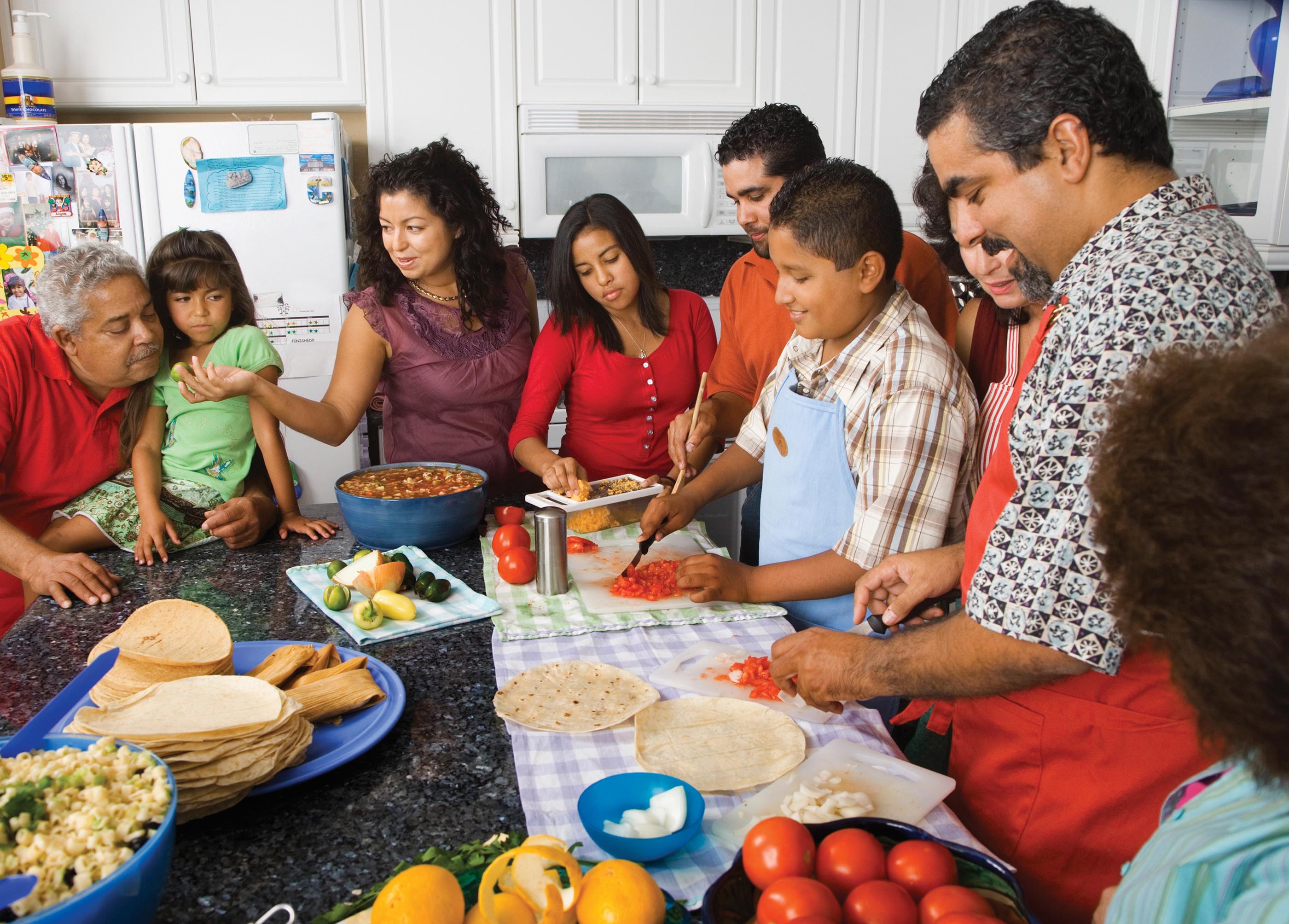 A boy and a man, wearing aprons, chop tomatoes on a table. The table is laden with tortillas, vegetable, fruits, choppers, and plates. A girl grates vegetables beside them, and a few people are gathered around the table.