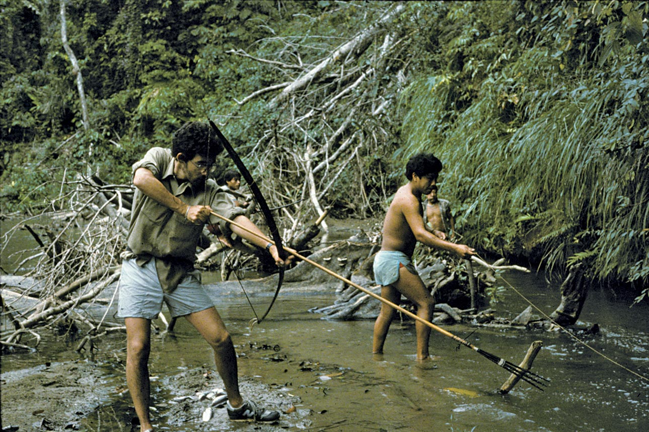 Lawrence Sugiyama stands in a swamp, carrying a bow and arrow and takes aim, while another man is holding a rope.