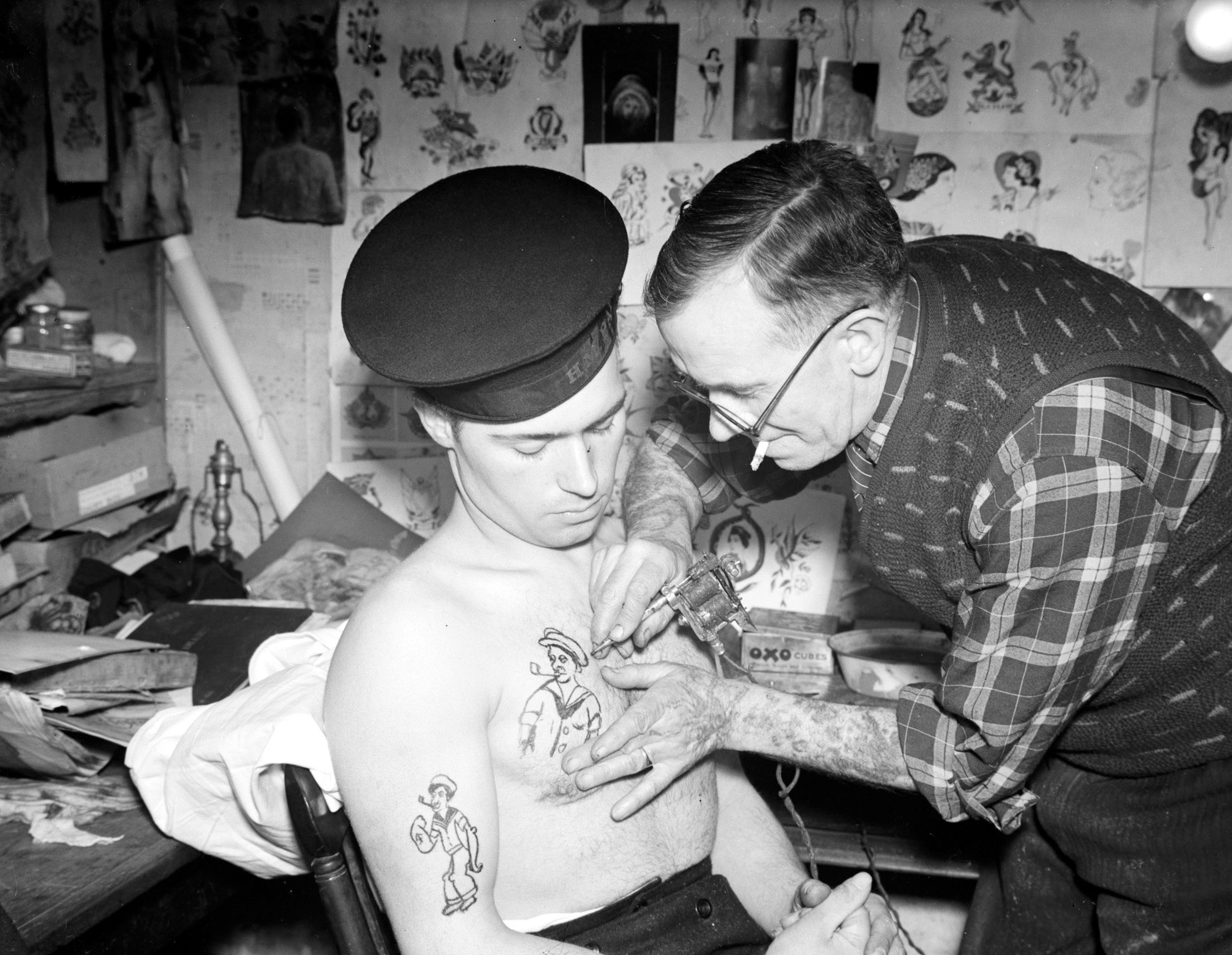 A black-and-white photo shows a tattoo artist with a cigarette in his mouth and glasses over his eyes. He draws a tattoo on the left side chest of a sailor. The sailor wears a hat, sits with a bare chest, and tries to look at the tattoo.