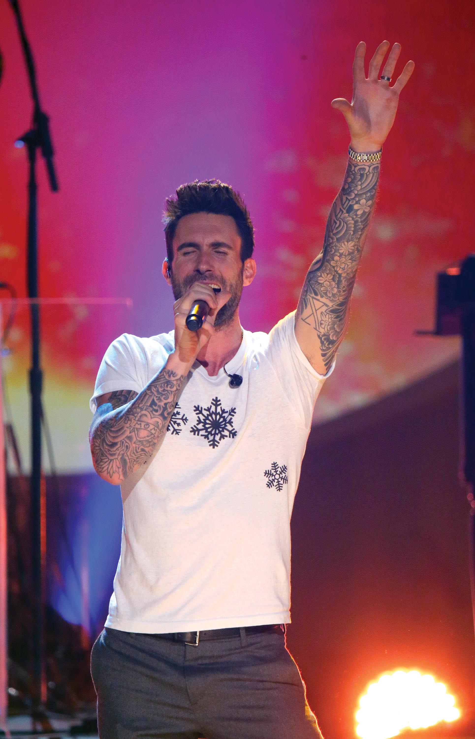 Lead vocalist, Adam Levine, sings while holding a mike in his right hand and raising his left hand to the audience. The vocalist has tattoos on both his arms. He wears a white t-shirt with snowflakes embroidered on it.