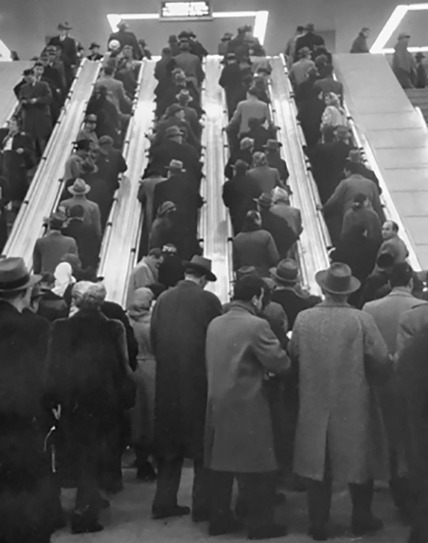 A black-and-white photo of a crowded area. The photo shows people, in organized rows, using the escalators.