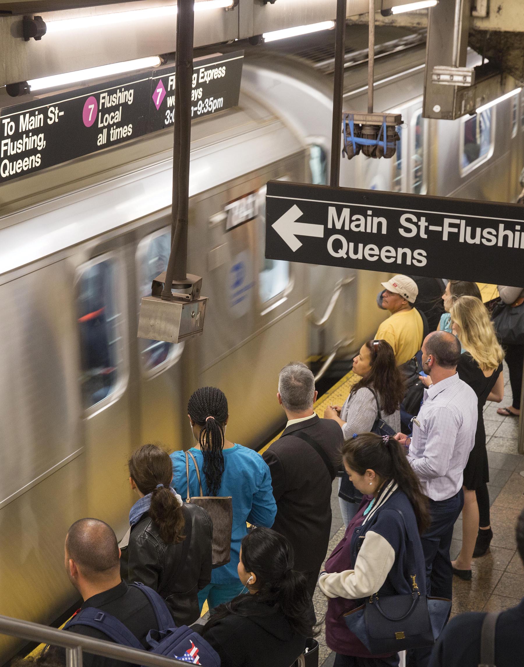 A photo shows the scene at a metro station where people are standing near the metro tracks and the train is in motion. A board indicates the destination of the metro train, which reads, Main Street-Flushing, Queens. Another board indicates some more information about the train.