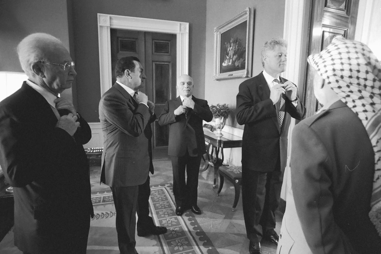 Bill Clinton, Yitzhak Rabin, Hosni Mubarak, and King Hussein of Jordan standing in a room, wearing formal suits, and adjusting their ties. Another person with his back to the camera wears a keffiyeh.