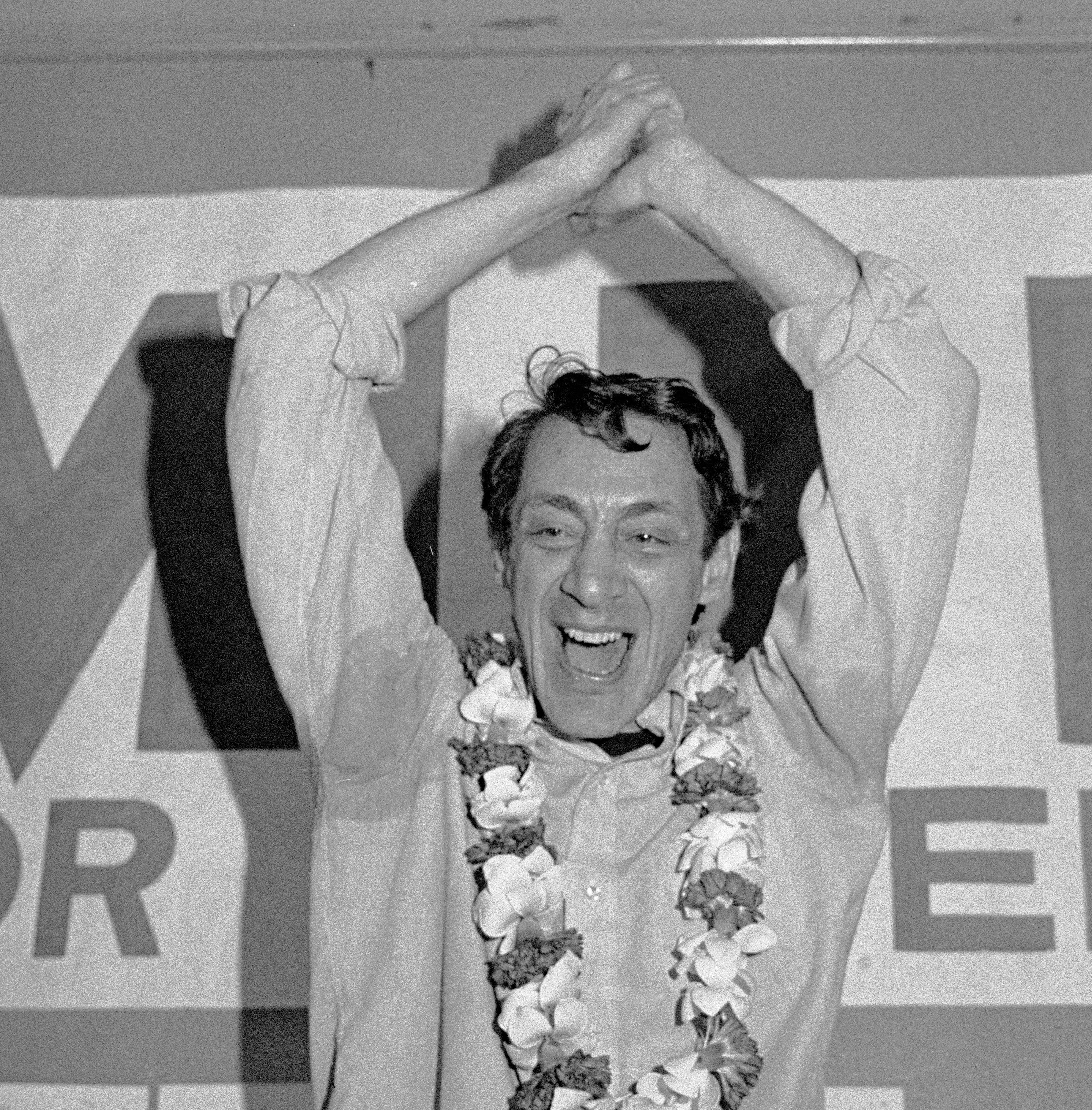 A black-and-white photo of Harvey Milk wearing a garland and smiling victoriously with his hands together in the air.