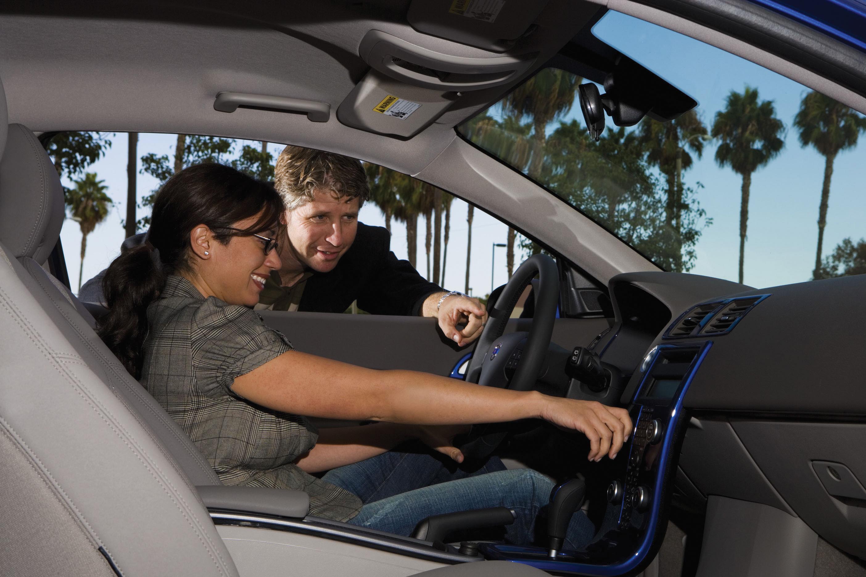 A young lady smiles and sits on the driving seat of a car, while a man guides her from the window of that seat.