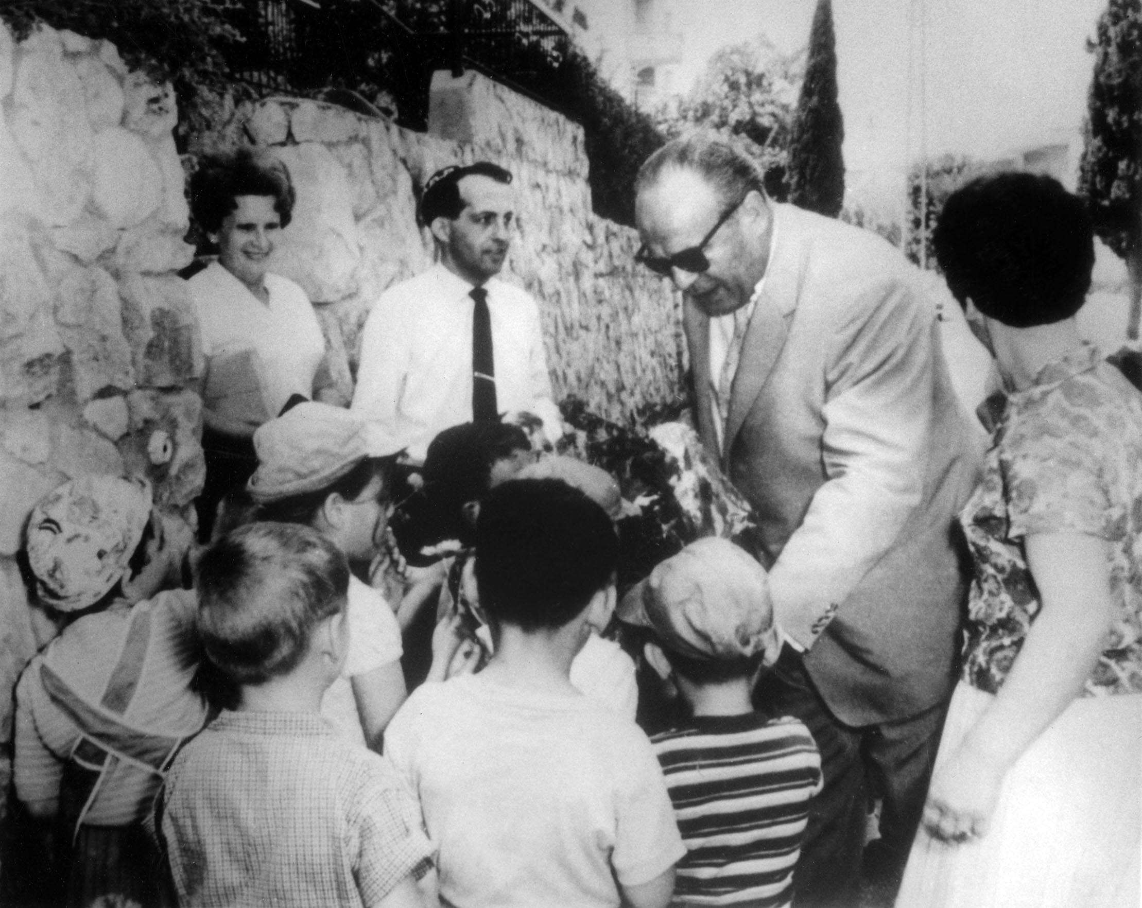 Oskar Schindler bends and talks with a group of Jewish kids in Tel Aviv. Two women and a man standing with them.