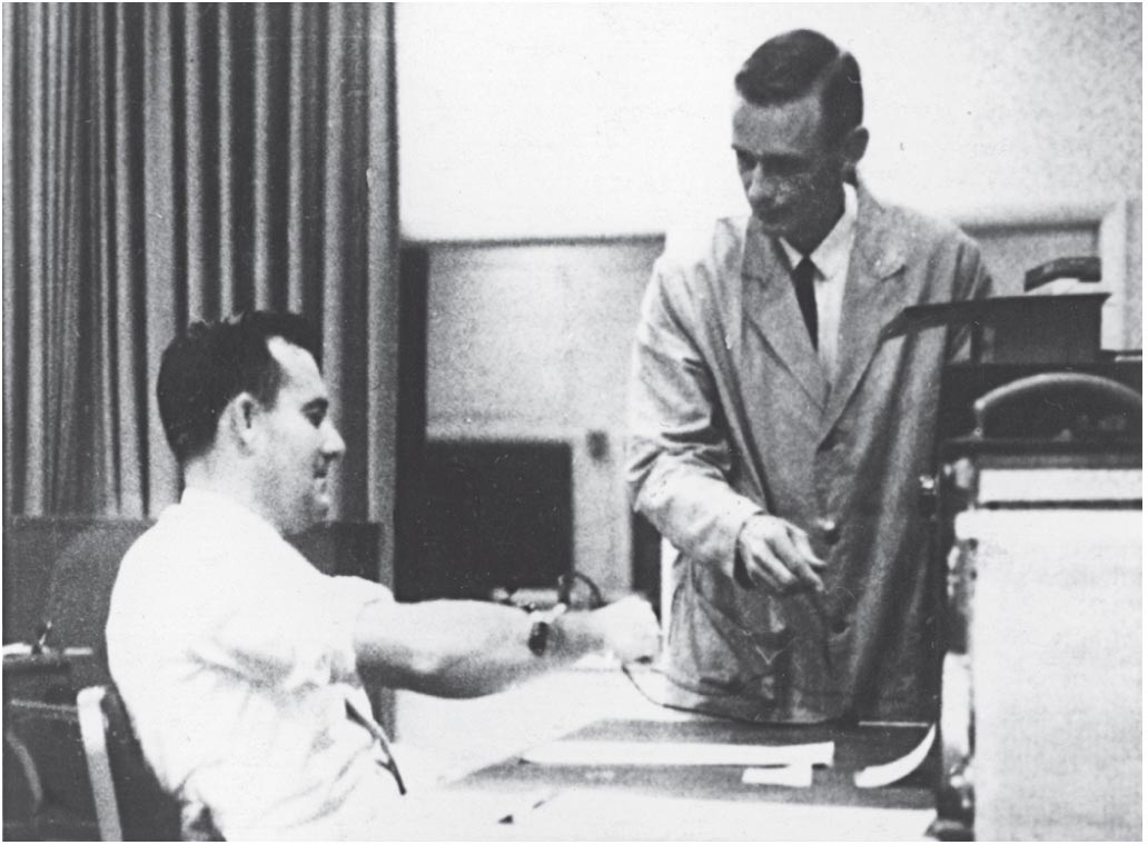 A participant sits on a chair. A man standing on his side attaches some wires to his right arm, that are connected to a shock generator.