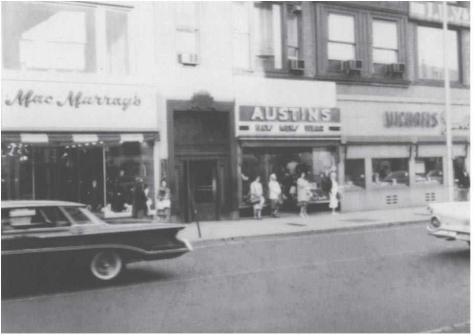 A black-and-white photo of a downtown area with various shops.