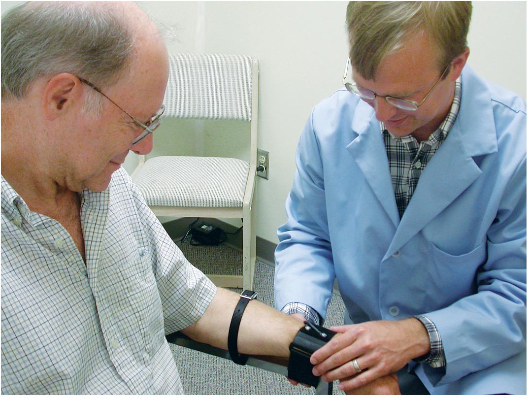 A photo shows two persons in a room where one person wearing a blue laboratory coat has tied the other person’s left arm with the chair. He fixes a machine on his left wrist.