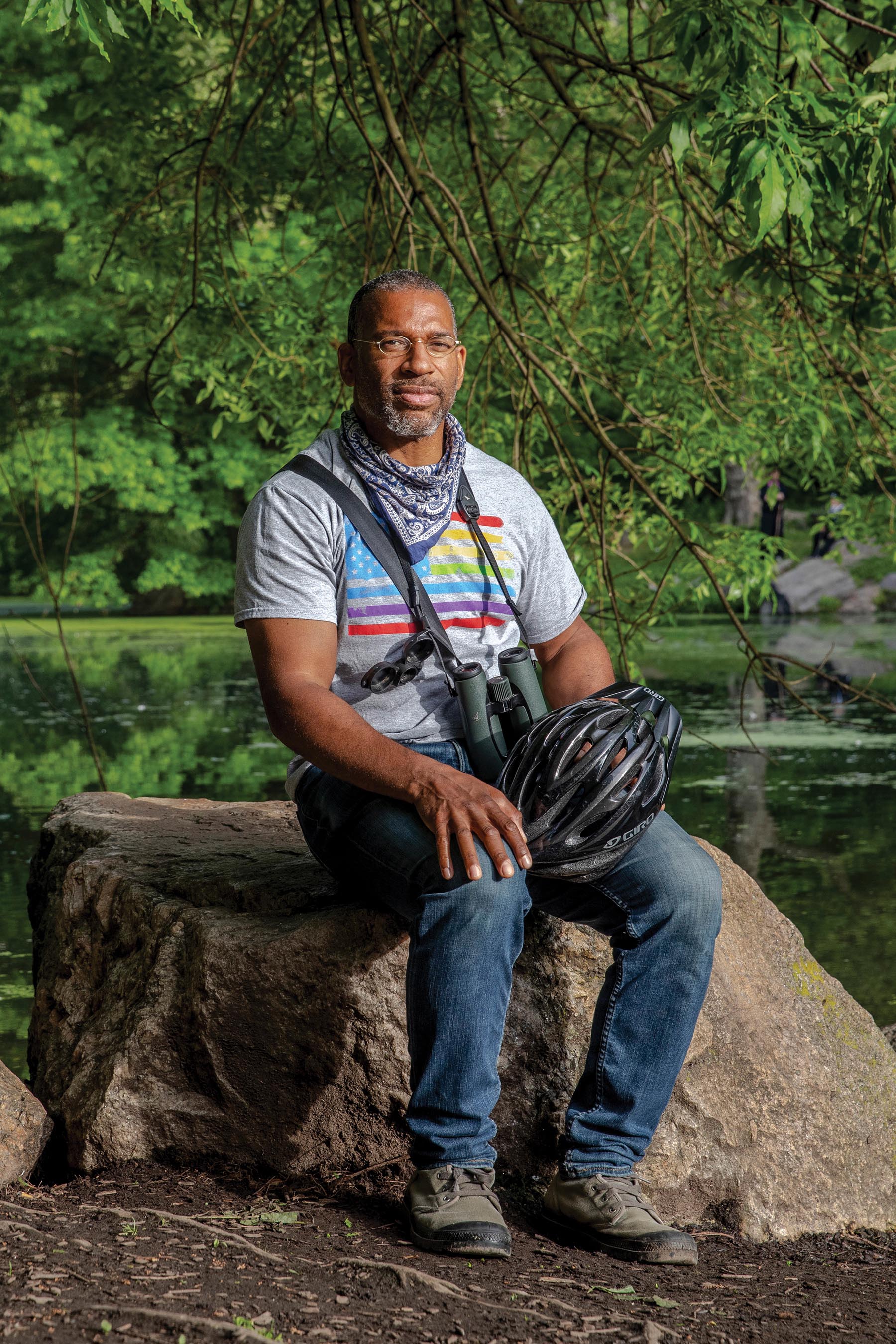 Christian Cooper sitting on a rock in a park, with a pair of binoculars hung around his neck.