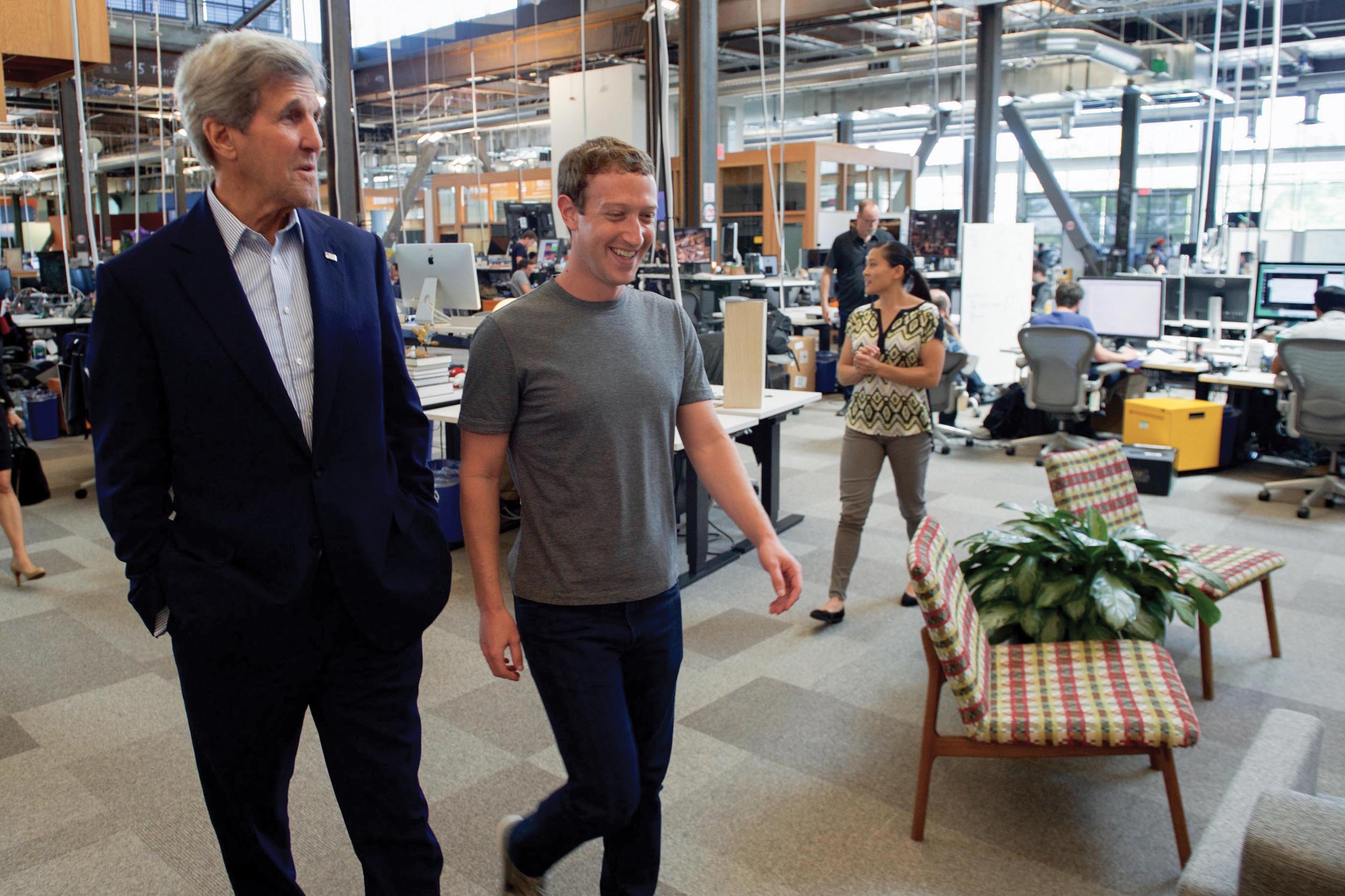 Mark Zuckerberg and John Kerry walking in an office and conversing.