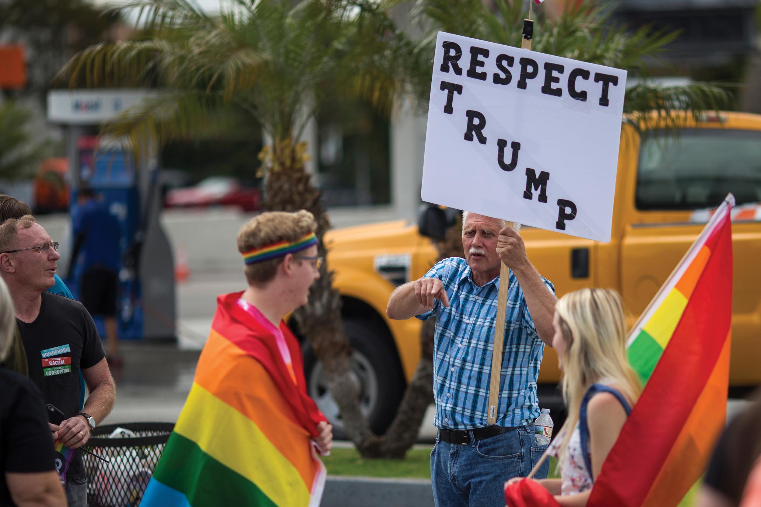 An elderly man holding a placard that reads, “Respect Trump.” He points his finger at a man who has a colorful flag wrapped around him.