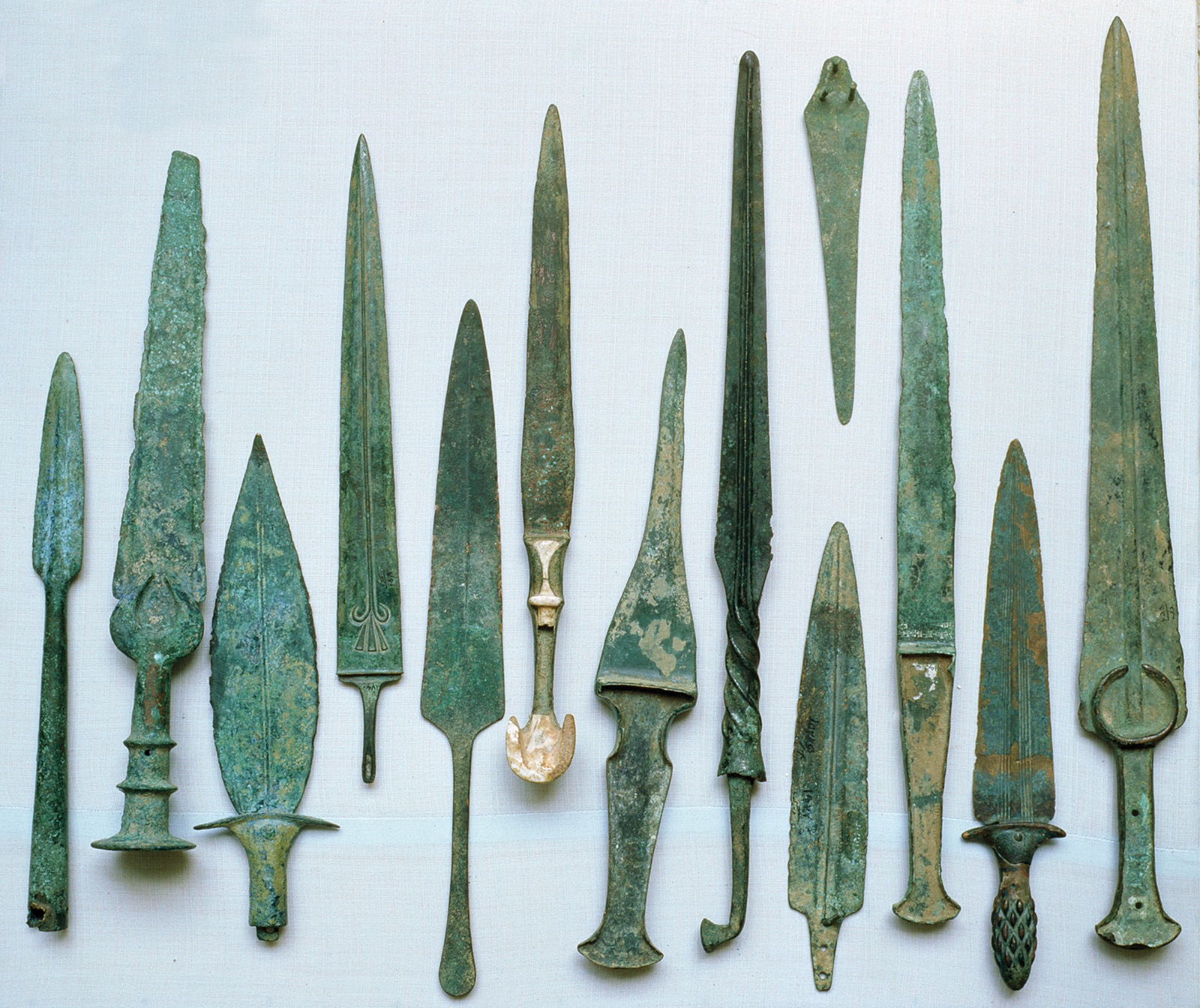 A photo shows thirteen rusted bronze knives of various forms and sizes, some of which have elaborate designs.