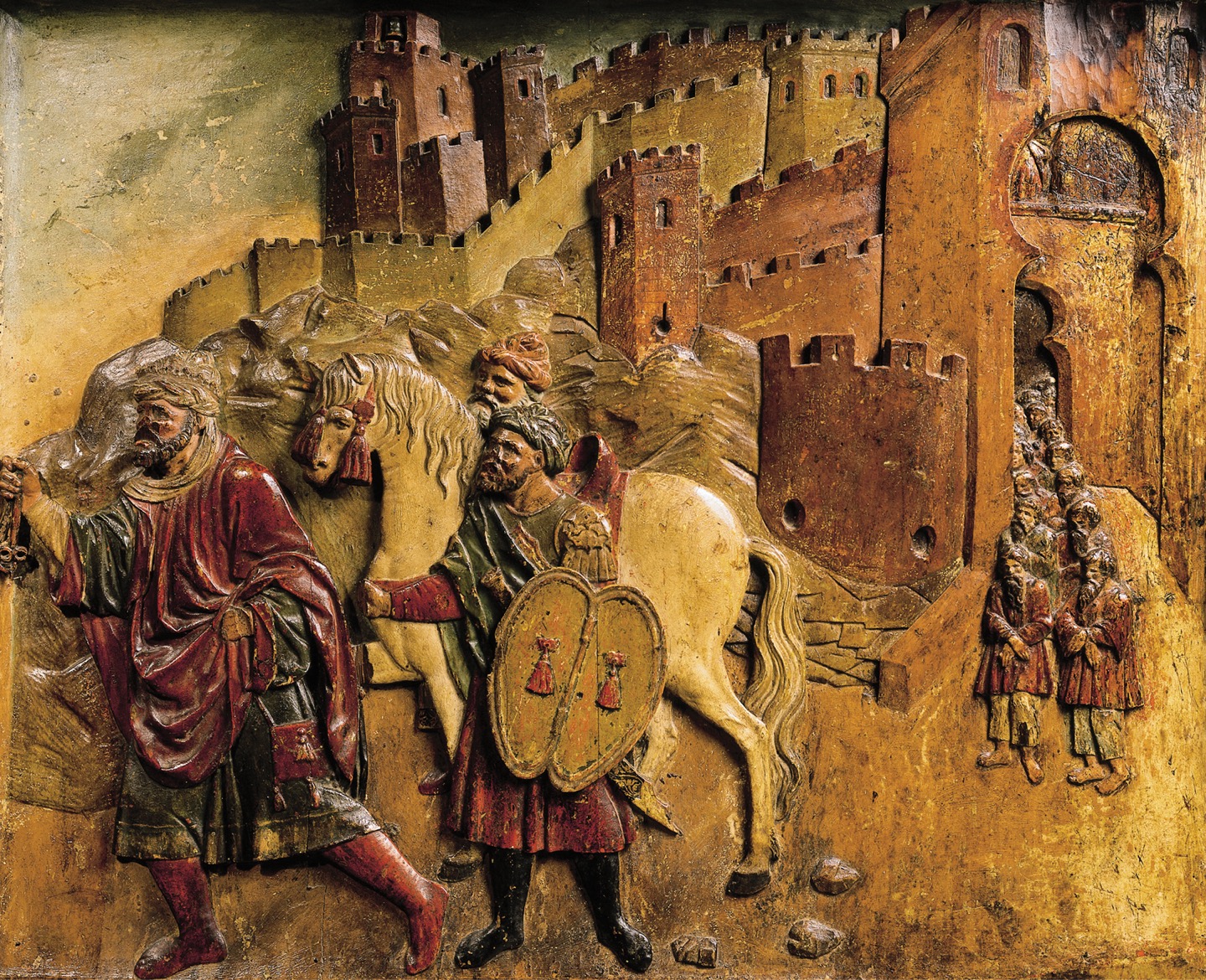A wooden sculpture shows a man leading two men, a horse, and men in two straight lines, away from the entrance to a fortress.