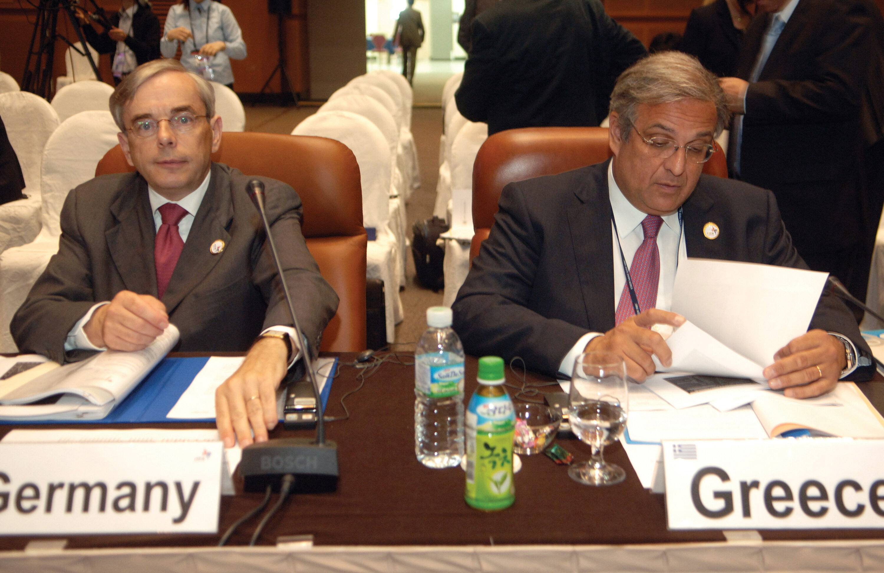 Two men in suits sitting at desks, one representing Germany and the other Greece. There are many people behind them in the large room.