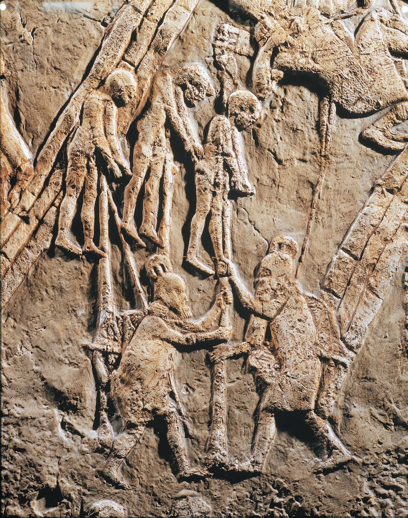 Stone carving of two soldiers holding a sharp-tipped pole figure and a dead, pierced, hanging man at the top of the pole. There are other two dead, hanging men.