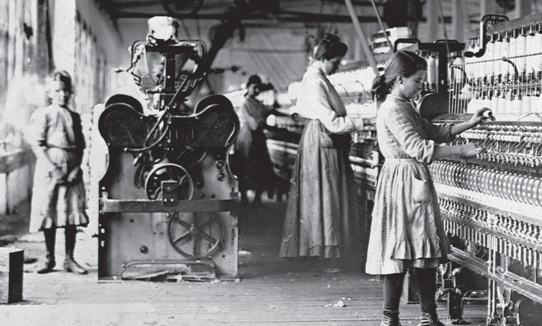 In a black and white photo, a child and several women work at a large machine inside a mill building.