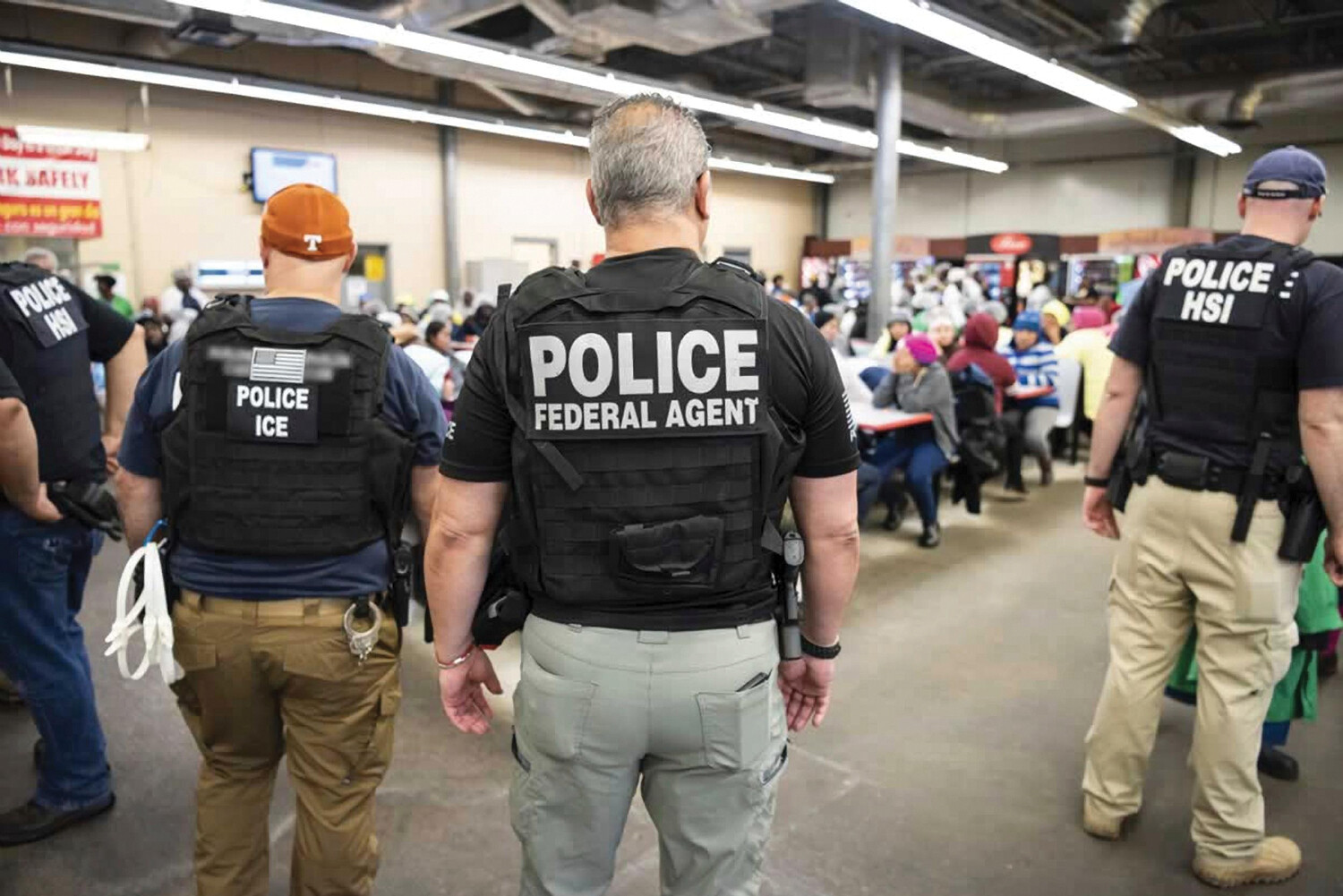 Four police officers in bulletproof vests watch a group of people seated at tables.