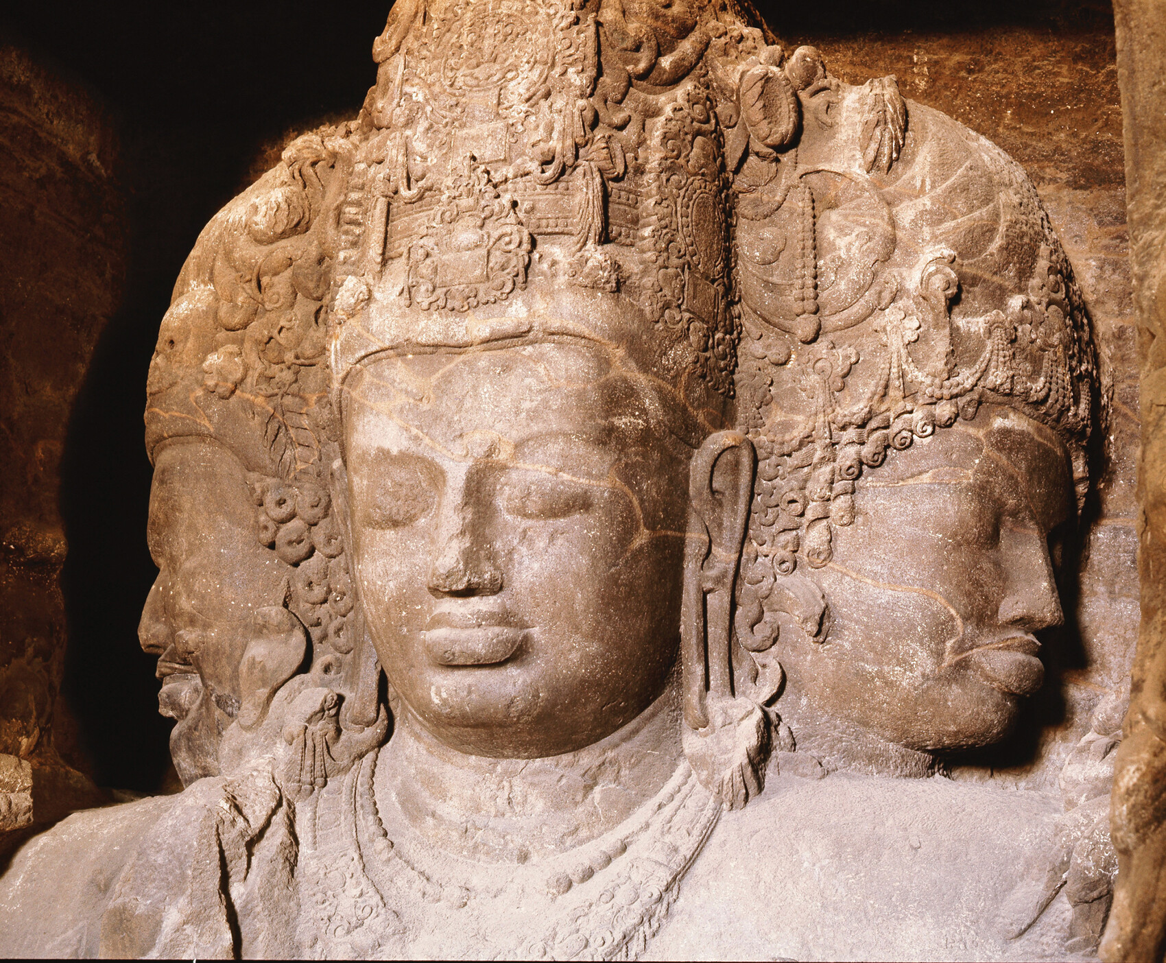A massive stone sculpture carved into a cave wall shows a three-headed Hindu deity.