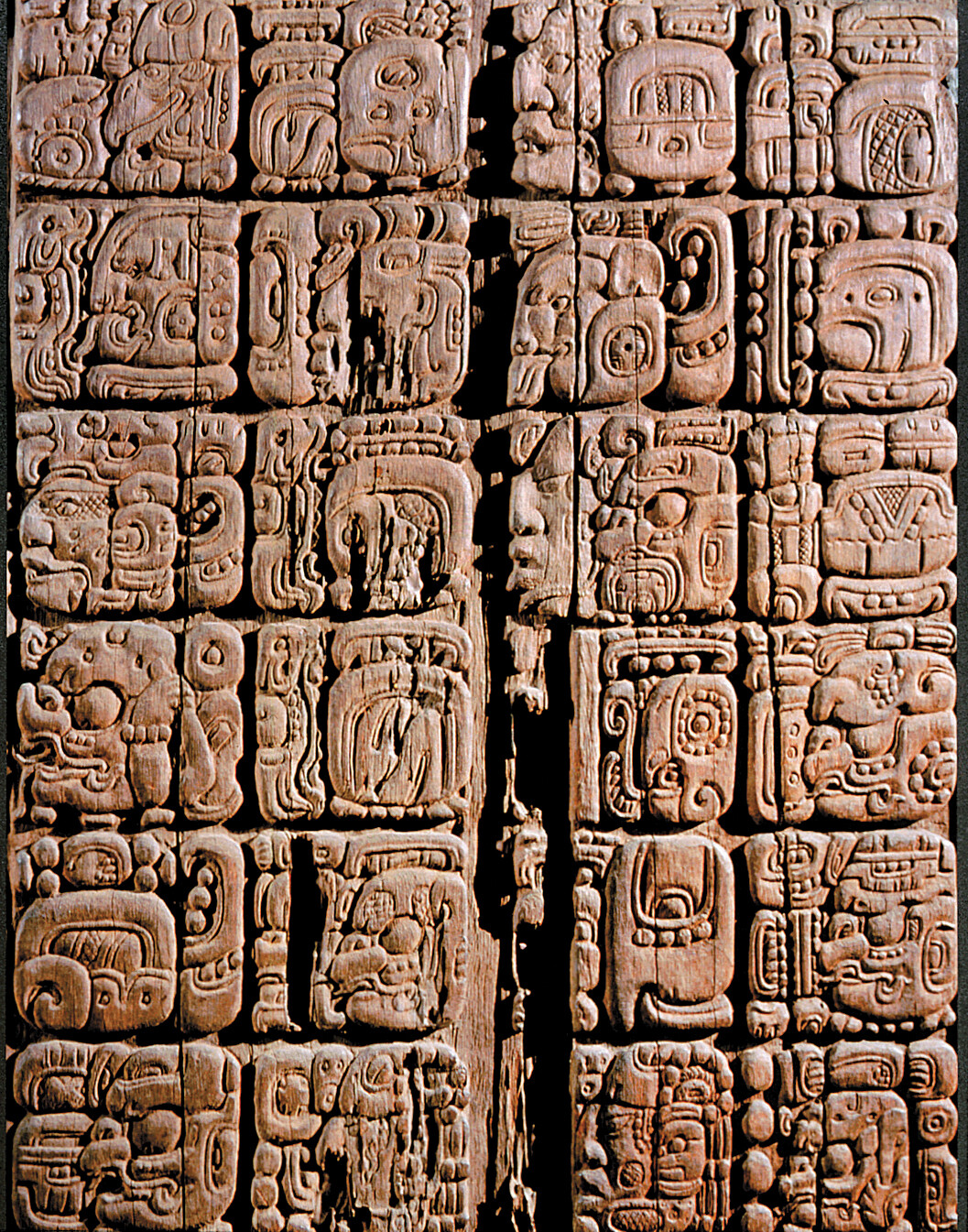 A close view of a carved wooden tablet shows rows of square panels filled with detailed glyphs and small carved images.